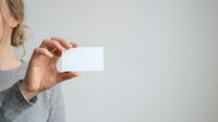 Woman with blonde hair wearing gray sweater holding blank white business card or mockup template against neutral background for branding and corporate identity projects.