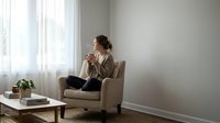 Young woman relaxing in modern armchair by window drinking coffee in minimalist living room interior with natural light and cozy atmosphere.