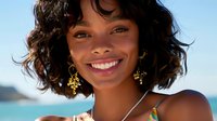 Young mixed-race woman with curly hair smiling at tropical beach wearing gold earrings and colorful swimsuit on sunny vacation day.