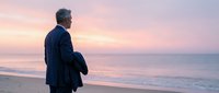 Mature businessman in suit standing on beach at sunset contemplating future decisions with ocean waves and colorful sky backdrop for business success concepts.