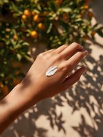 Female hand with white moisturizing cream against citrus tree background with orange fruits and leaf shadows creating natural summer skincare composition.