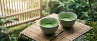 Two ceramic bowls filled with green matcha tea on bamboo mat in peaceful Japanese garden setting with natural lighting and traditional fence background.