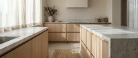 Modern kitchen with light wood cabinetry, marble countertops, and minimalist design. Bright natural light from windows illuminates the clean, contemporary space with neutral tones.
