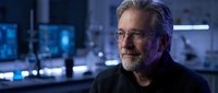 Mature man with gray hair and beard wearing glasses in modern tech office environment with blue-lit screens and digital displays at night.