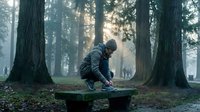 Man in dark athletic wear crouching on wooden bench in misty forest with tall trees, contemplative mood, suitable for wellness, fitness, and mental health concepts.