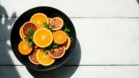 Sliced citrus fruits including oranges and grapefruits arranged on black plate with mint leaves, shot from above on white wooden surface for food styling and culinary content.