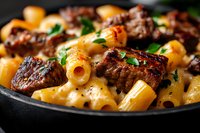 Rigatoni pasta with grilled beef chunks in creamy cheese sauce garnished with fresh parsley, served in black plate. Close up macro food photography.