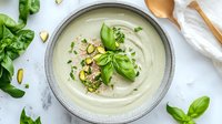 Creamy cauliflower soup garnished with fresh basil leaves, chopped pistachios, and herbs in gray ceramic bowl on white marble background. Food styling.