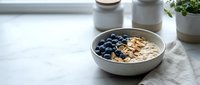 Oatmeal bowl with blueberries and granola on marble countertop near window with potted plants and ceramic containers for breakfast and healthy eating.