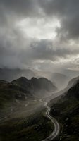 Dramatic stormy clouds over winding mountain road through rugged highland valley with moody atmospheric lighting and misty peaks in remote wilderness landscape.