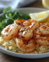 Grilled seasoned shrimp served on orzo pasta with fresh herbs, black pepper, and lemon wedges garnish on white plate. Close up view of seafood dinner.