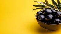 Black olives in golden bowl with olive branch on bright yellow background, suitable for food photography, culinary content, and Mediterranean cuisine marketing materials.