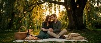 Couple enjoying romantic picnic under large tree with golden sunlight filtering through foliage, sitting on blanket with basket and wine for outdoor dating and relationship concepts.