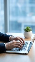 Professional businessman hands typing on laptop computer at modern office desk with succulent plant and blurred city view background for remote work concepts.