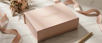 Elegant blush pink thank you gift box with gold lettering on marble surface, surrounded by satin ribbon and flowers, perfect for weddings and celebrations.