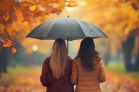 Two women with long hair sharing umbrella while walking in autumn park. Golden fall foliage and bokeh create romantic atmosphere on rainy day. Back view portrait.