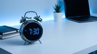 Retro alarm clock with digital blue LED display showing 7:30 AM on a white desk with laptop and plant in cool blue light.