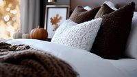 Cozy autumn bedroom decor with knitted brown pillows, soft white bedding, and pumpkin decoration against blurred warm lights creating seasonal ambiance.