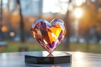 Crystal heart prism refracting sunlight creates rainbow colors and warm golden glow, displayed on reflective surface against blurred natural background at sunset.