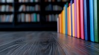 Colorful book spines lined up on dark wooden table with blurred library background, creating depth of field effect with selective focus on vibrant book edges.