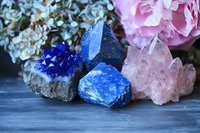 Raw blue lapis lazuli and pink rose quartz crystal clusters with hydrangea and peony flowers on dark textured background for spiritual healing practices.