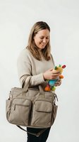 Woman holding colorful toy while wearing beige diaper bag with multiple pockets. Caucasian female, mid-thirties, smiling, dressed in cream sweater and black pants, carrying practical parenting accessory for babies and toddlers.