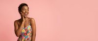 Happy young African American woman in colorful floral dress talking on smartphone against pink background, expressing joy and excitement during phone call.