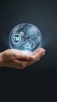 Hand holding glowing sphere with trademark symbols, brain, lightbulb and gears representing intellectual property protection and innovation concepts.