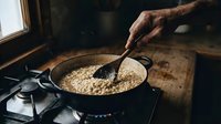 Mature male hand stirring cooked grains in cast iron skillet on gas stove in rustic kitchen setting with natural window light and wooden surface.