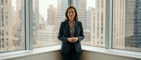 Professional businesswoman in dark suit standing confidently by office windows with urban cityscape background for corporate presentations and leadership content.