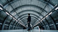 Silhouette of man in dark suit standing alone on empty train platform with symmetrical arched ceiling and fluorescent lighting, conveying solitude and urban commute atmosphere.