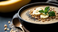 Creamy banana smoothie bowl topped with fresh banana slices, chia seeds, granola, and mint leaves garnish in black ceramic bowl on dark background.