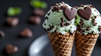 Mint chocolate chip ice cream scoops in waffle cones topped with heart-shaped chocolates and sprinkles against dark background, selective focus food photography.
