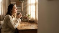 Young woman enjoying morning coffee in cozy kitchen with warm sunlight streaming through window creating peaceful domestic atmosphere for lifestyle content.