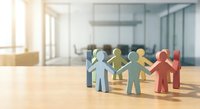 Colorful paper cutout figures holding hands in circle on wooden desk in modern office space representing teamwork, unity and collaboration concepts.