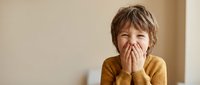 Happy young boy with tousled brown hair covering mouth while laughing in cozy mustard sweater against neutral background for family lifestyle content.