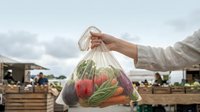 Farmer holding transparent plastic bag filled with fresh red peppers, green cucumbers, and ripe tomatoes at outdoor farmers market or agricultural harvest stand with blurred background.