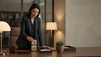 Professional businesswoman in dark suit standing at modern office desk with wooden blocks, warm lighting creates productive corporate workspace atmosphere.