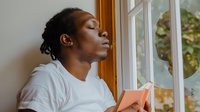 Young African American woman relaxing by window with book, enjoying peaceful reading moment in natural light for wellness and self-care concepts.