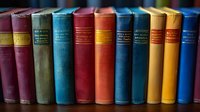 Row of vintage multicolored hardcover books standing on wooden shelf, showing aged cloth spines with gold lettering in classic library setting.