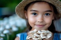 Young Asian girl in straw hat holding fresh mushrooms while smiling outdoors, with soft bokeh background of white flowers in garden setting.