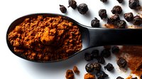Ground turmeric powder in black spoon with whole black peppercorns scattered on white surface, macro food photography showing vibrant orange spice texture.