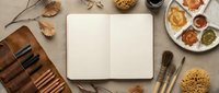 Open blank notebook surrounded by autumn art supplies including watercolor paints, brushes, and natural elements on beige canvas background for creative projects.