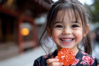 Cheerful Asian girl in traditional dress holding orange candy, showing bright genuine smile with dimples. Natural outdoor setting with soft bokeh background.