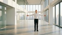 Professional businessman with arms outstretched standing in modern glass office lobby with city skyline view, corporate success and achievement concept.