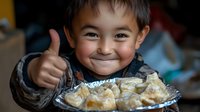 Young Asian boy showing thumbs up with excited smile while enjoying dumplings on aluminum plate, expressing joy and satisfaction during mealtime.