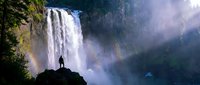 Silhouette of person standing on rock viewing majestic waterfall with rainbow mist in sunlight surrounded by lush forest landscape for adventure travel.
