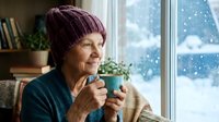 Senior Caucasian woman in burgundy knit hat enjoying hot beverage by snowy window. Cozy winter morning scene with books and plants creating warm atmosphere.