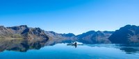 Serene alpine lake with mirror-like blue water reflecting snow-capped mountain peaks and forested slopes, featuring a lone boat for travel and adventure themes.