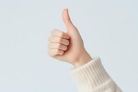 Female hand in white knitted sweater sleeve showing thumbs up gesture against light background. Positive feedback and approval concept.